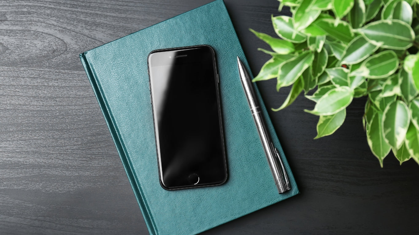 iPhone placed on a notebook beside a pen on a table, representing a guide on how to remove scratches from a phone screen.